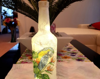 Puede incluir: Una botella de vidrio blanco con un tapón de corcho y un diseño de loro colorido. La botella está iluminada desde adentro, creando un brillo cálido.