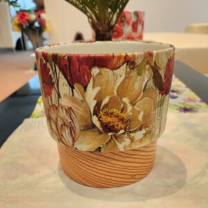 May include: A ceramic planter with a floral pattern in shades of red, yellow, and brown. The planter has a wooden base.