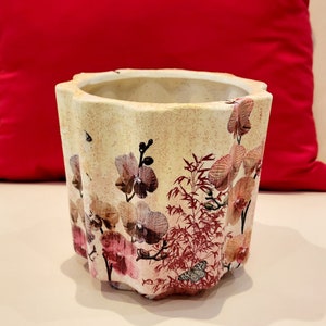 Puede incluir: Una maceta de cerámica blanca con un borde festoneado presenta un diseño floral de orquídeas rosas y marrones y tallos de bambú rojos. La maceta tiene un aspecto desgastado y vintage.