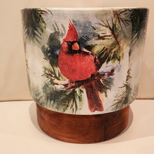 May include: A ceramic planter with a winter scene featuring a red cardinal perched on a branch. The planter has a wooden base.