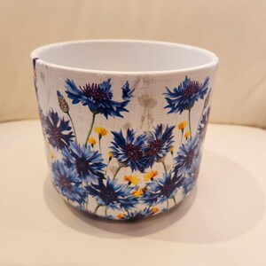May include: White ceramic planter with a blue and yellow floral pattern. The pattern features blue cornflowers and yellow flowers.