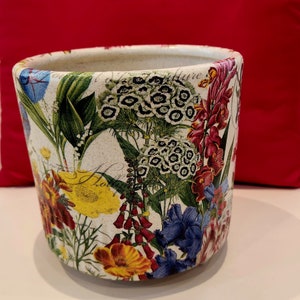 May include: A white ceramic flower pot with a floral pattern featuring various flowers in shades of red, yellow, blue, and green. The pot has a textured surface and a slightly rounded shape.