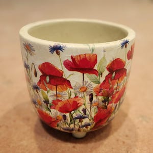 May include: A cylindrical ceramic planter with a cream-coloured base featuring a vibrant botanical print of red poppies, blue cornflowers, and white daisies. The vessel has a smooth rim, a deep interior, and is supported by small, subtle feet.