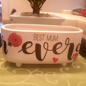 May include: White ceramic planter with the words "Best Mum Ever" in black script. The planter has a red heart and two red roses on the side. It has three small feet on the bottom.