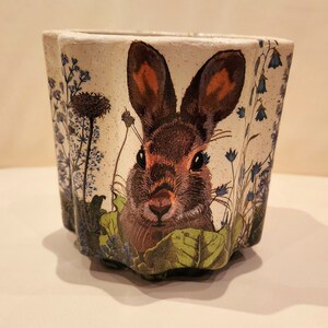 May include: A white ceramic planter featuring a brown rabbit face and blue flowers printed on the front.