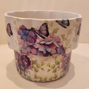 May include: A cylindrical planter with a scalloped top edge, featuring a decorative print of purple and blue hydrangea flowers, green foliage, and several butterflies in shades of blue and purple against a cream-coloured background.