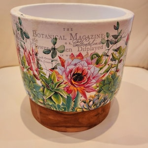 May include: A ceramic plant pot with a botanical design. The pot has a white rim, a floral print with pink and green succulents, and a wooden base. The text on the pot reads "THE BOTANICAL MAGAZINE".