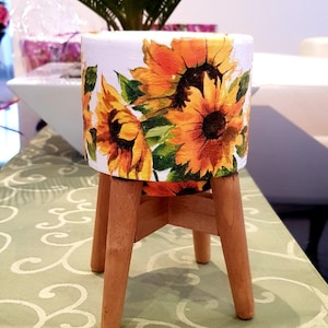 May include: A white ceramic planter with a sunflower design. The planter has three wooden legs.