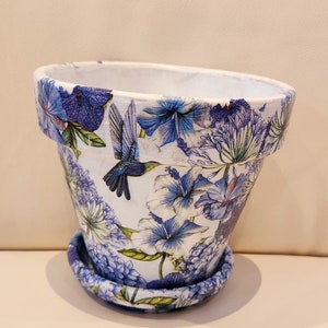 May include: A white ceramic flower pot with a blue and white floral design featuring hummingbirds. The pot has a matching saucer.