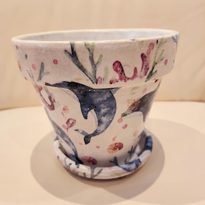 Peut inclure: Pot en céramique blanche avec un motif de dauphin bleu et de corail. Le pot a une soucoupe assortie.
