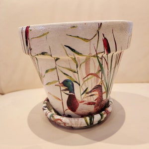 May include: A white ceramic planter with a duck and reeds design. The planter has a matching saucer.