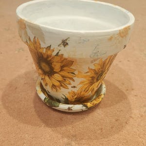 May include: A white ceramic flower pot with a matching saucer. The pot is decorated with large yellow sunflowers and small bees. The pot has a wide opening and a slightly tapered shape. The saucer is round and has a similar sunflower design.