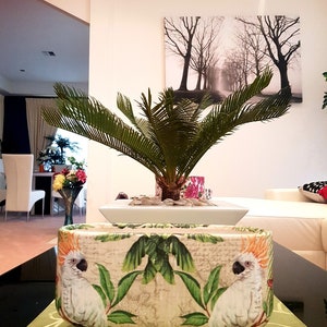 May include: A white ceramic planter with a tropical design featuring two white cockatoos with orange crests. The planter is sitting on a green table runner with a green plant in the background.