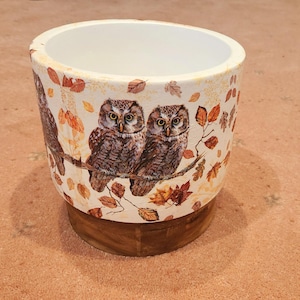 May include: A cylindrical ceramic planter with a white interior and a decorative exterior. The exterior features two owls perched on a branch, surrounded by autumn leaves. The planter sits on a wooden base.