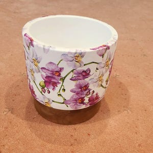May include: A white ceramic plant pot decorated with a floral orchid pattern in purple and white. It rests on a round wooden base. The inside of the pot is white.