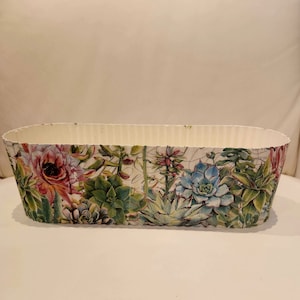 Puede incluir: Maceta ovalada blanca con un diseño floral que presenta suculentas y flores en tonos verdes, rosas y azules.