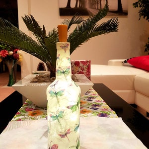 May include: A clear glass bottle with a cork stopper and a white background decorated with colourful dragonfly designs. The bottle is illuminated from within, creating a warm glow.