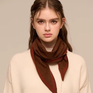 May include: A brown scarf with a red trim is worn around the neck. The scarf is made of a soft material and is worn over a cream-coloured jumper. The scarf's ends are pointed.