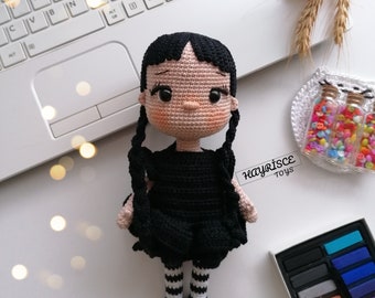 May include: A crocheted doll with black hair, a black dress, and white and black striped stockings. The doll is sitting on a white surface with a laptop and other craft supplies in the background. The text "HAYRİSCE Toys" is visible in the image.