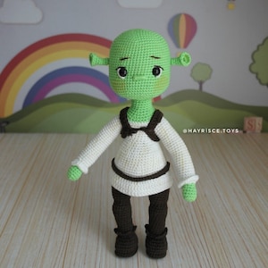 May include: A green crocheted plush toy of Shrek, a character from the movie Shrek, wearing a white shirt and brown pants. The toy has a friendly expression and is standing on a wooden surface. The text "@HAYRISCE.TOYS" is visible in the background.