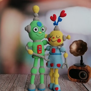 May include: Two crocheted robot toys, one green and one yellow, standing side by side. The green robot has a blue and red button detail on its chest. The yellow robot has a blue dress with red hearts and a red and blue flower on its head.