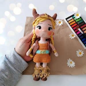 May include: A handmade crochet doll in the shape of a Viking girl. The doll has blonde braids, a horned helmet, and an orange dress with a blue belt. The doll is wearing brown boots with fringe.