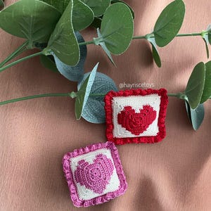 Pode incluir: Duas pequenas almofadas quadradas de crochê com desenhos de coração. Uma almofada apresenta um coração vermelho sobre um fundo branco com uma borda vermelha. A outra tem um coração rosa com uma borda rosa. Folhas verdes no fundo.