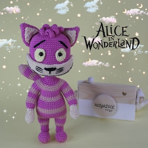 May include: A crocheted purple and white striped cat with yellow eyes, a black nose, and a black mustache. The cat is standing on a light pink surface with a wooden camera in the background. The camera has a white label that says "Hayrisce Toys". The text "Alice in Wonderland" is written in black on a light pink background.