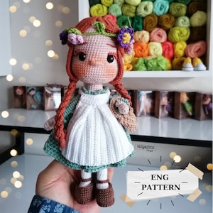 Puede incluir: Muñeca de crochet con pelo rojo, vestido verde, delantal blanco y zapatos marrones. La muñeca lleva una cesta pequeña de crochet. La muñeca lleva una corona de flores.
