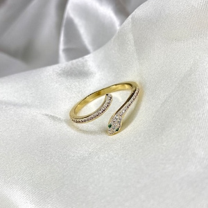 May include: A gold snake ring with a wrap-around design. The ring is adorned with clear and pink gemstones along the body and two green gemstones for the eyes. The ring is set against a white, silky background.