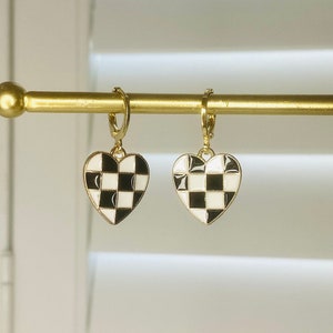 May include: Gold-coloured hoop earrings with heart-shaped pendants. Each pendant has a black and white chequered pattern, outlined in gold. The earrings are displayed on a gold bar against a blurred background.