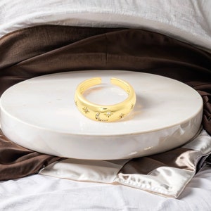 May include: A gold-coloured adjustable ring with a celestial design. The ring features a crescent moon and star pattern, with small clear stones. The ring is displayed on a white, round surface.