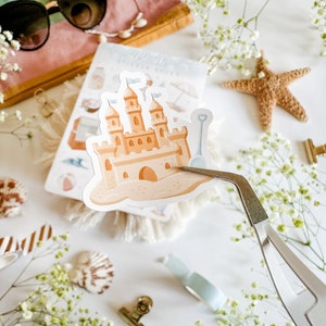 May include: A sticker of a sandcastle with a blue bucket and shovel on a white background. The sticker is being held by tweezers.