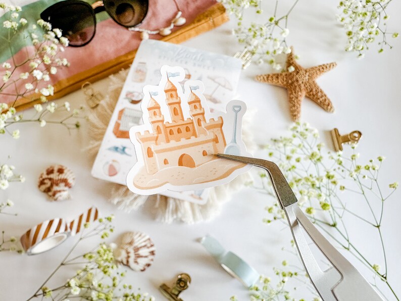 May include: A sticker of a sandcastle with a blue bucket and shovel on a white background. The sticker is being held by tweezers.