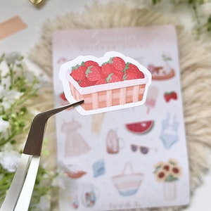 May include: A sticker of a wicker basket filled with red strawberries. The sticker is on a white background with other stickers of summer-themed items.