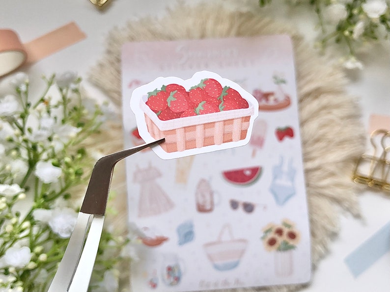 May include: A sticker of a wicker basket filled with red strawberries. The sticker is on a white background with other stickers of summer-themed items.