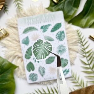 May include: A sheet of tropical leaf stickers with a variety of green leaves, including monstera leaves, palm leaves, and ferns. The stickers are on a white background with the text "Tropical Leaves Sticker Sheet" printed at the top. A pair of tweezers is holding one of the monstera leaf stickers.