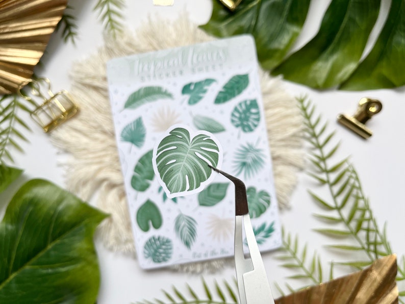May include: A sheet of tropical leaf stickers with a variety of green leaves, including monstera leaves, palm leaves, and ferns. The stickers are on a white background with the text "Tropical Leaves Sticker Sheet" printed at the top. A pair of tweezers is holding one of the monstera leaf stickers.