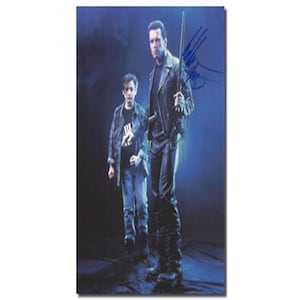 May include: A black and white photo of two people, one standing and one sitting. The person standing is wearing a black leather jacket and black pants. The person sitting is wearing a black shirt and blue jeans. The photo is signed by Arnold Schwarzenegger.