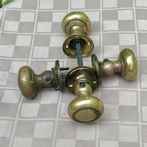 May include: A set of three brass door knobs with round handles and circular mounting plates. Each knob features a central metal spindle connecting the hardware components, showcasing a polished metallic finish with visible signs of age and wear.