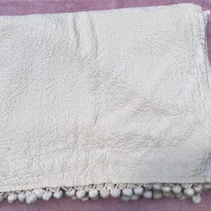 May include: Off-white quilted throw blanket with a textured surface and a row of white pom-poms along the bottom edge. The blanket is folded and appears soft and cozy, suitable for a bed or sofa.