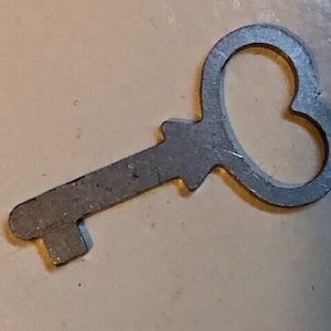 May include: A vintage-style metal key with a heart-shaped handle. The key is silver-colored and has a rectangular bit. The key is lying on a light beige surface.