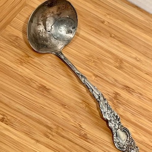 May include: An antique silver ladle with an ornate handle and a round bowl. The ladle is tarnished, showing signs of age and use. The handle features detailed floral and scrollwork designs, adding to its vintage charm.