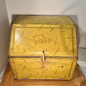 May include: Yellow metal cake carrier with a hinged lid and a latch closure. The carrier has a distressed finish with a pattern of intersecting lines. The word "Cake" is printed on the front.