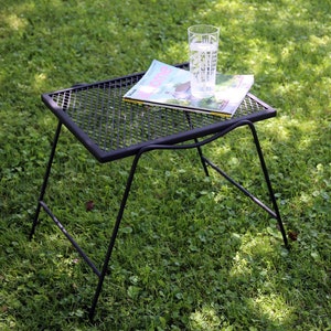 May include: A black metal side table with a rectangular mesh top and four thin angled legs. Resting on the table is a magazine with the word home visible on the cover and a clear glass tumbler featuring a white botanical leaf pattern design.