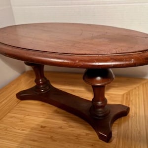 May include: A small, antique wooden table with a round top and a dark brown finish. The table has a pedestal base with three legs. The table is approximately 24 inches in diameter and 20 inches tall.