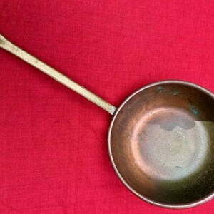 May include: An antique copper pan with a long, brass handle. The pan has a round bowl shape and is set against a red background. The handle has a hole at the end for hanging.