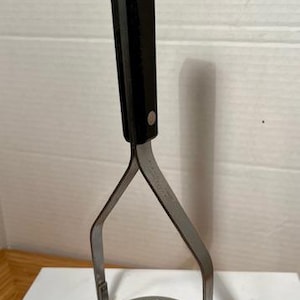 May include: A potato masher with a black handle and a stainless steel mashing head. The handle is rectangular and has a hole at the top. The mashing head is round with multiple holes.