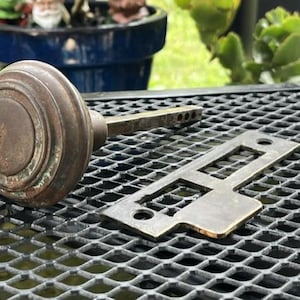 May include: A vintage round metal door knob with a weathered brown patina and a long square spindle, accompanied by a flat rectangular metal strike plate featuring two circular screw holes and a central rectangular cutout for a latch mechanism.