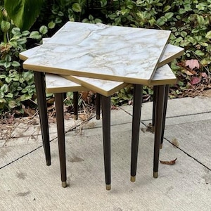 May include: A set of three nesting tables with square, marble-look tops and dark brown legs. The table edges are trimmed with gold-colored metal. The tables are arranged in a stacked configuration, showcasing their varying sizes and design.